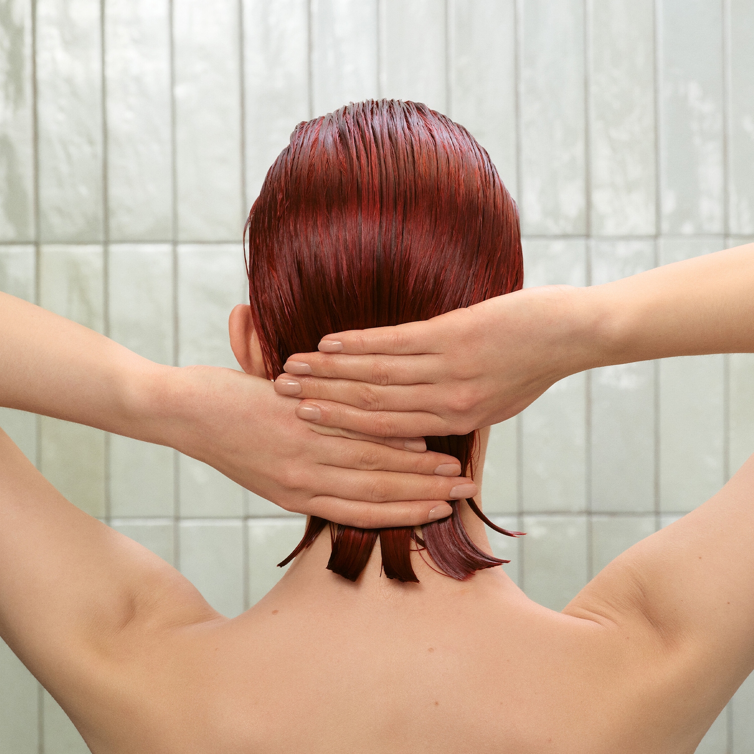 Vista posteriore di una donna con i capelli rosso scuro bagnati, le mani incrociate dietro la nuca, su sfondo di piastrelle bianche.
