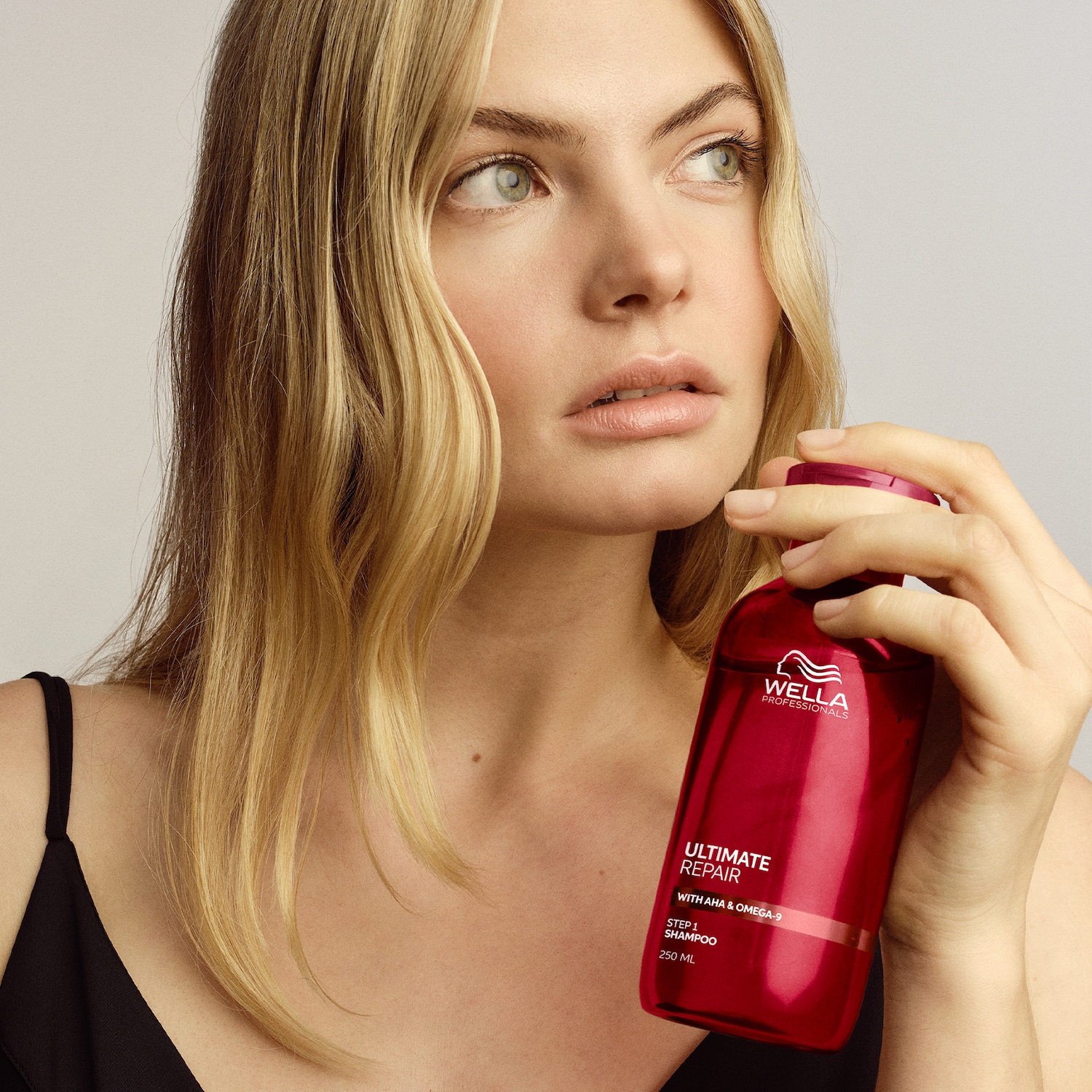 Una donna bionda tiene in mano un flacone rosso di shampoo Wella Professionals Ultimate Repair Step 1 con AHA e Omega-9, 250 ml.