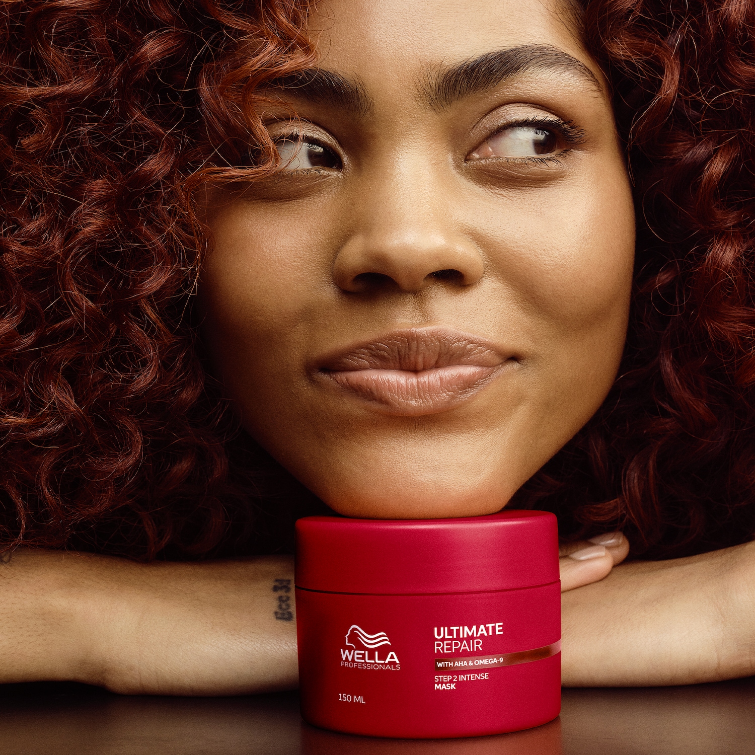 Primo piano di una donna con capelli ricci rossi appoggiata a un barattolo rosso di Wella Professionals Ultimate Repair Step 2 Intense Mask, che promette riparazione con AHA e Omega-9.