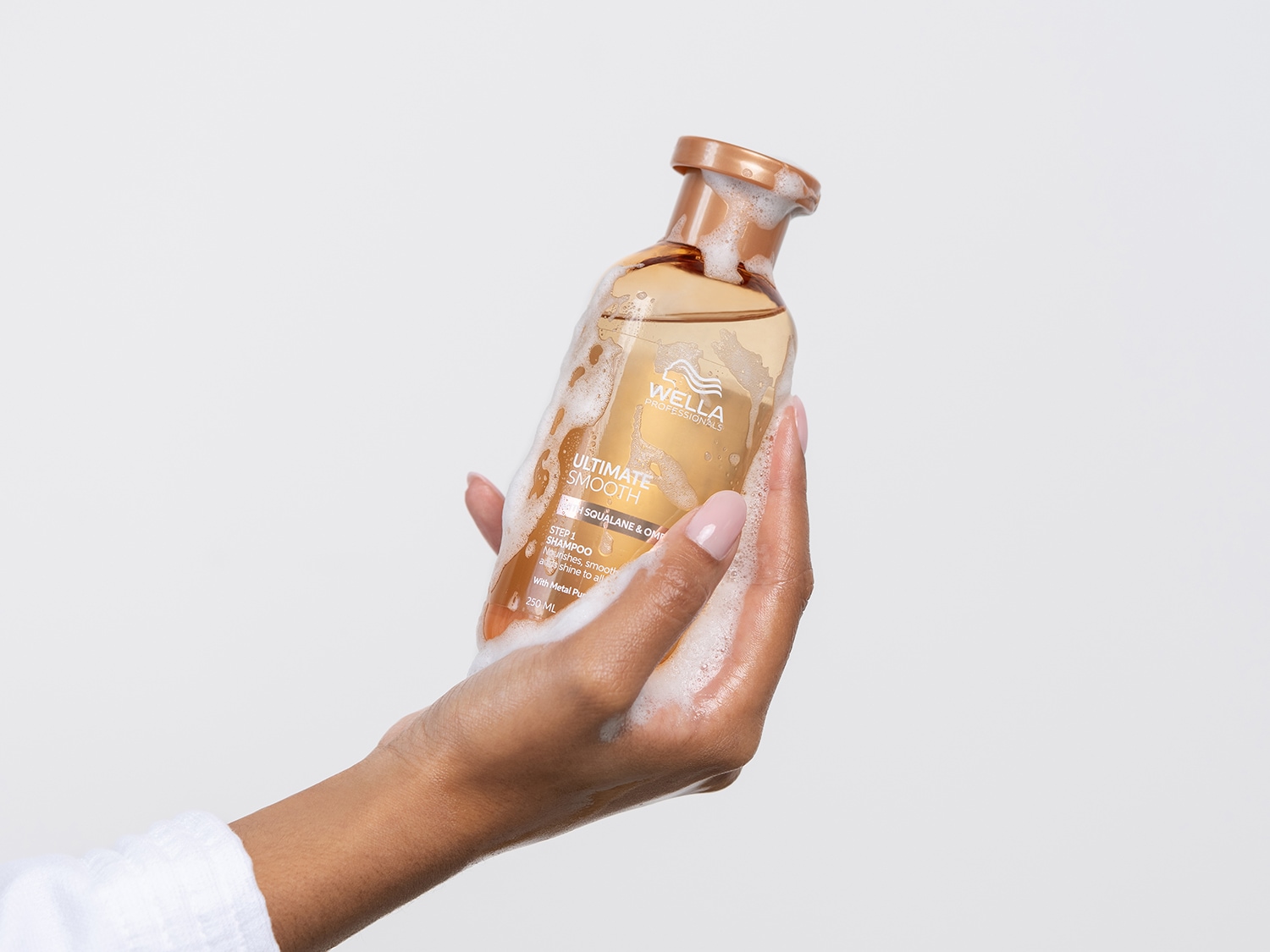 Una mano tiene una bottiglia di shampoo Wella Professionals Ultimate Smooth Step 1, con schiuma bianca che indica la sua consistenza nutriente e lisciante per capelli luminosi.
