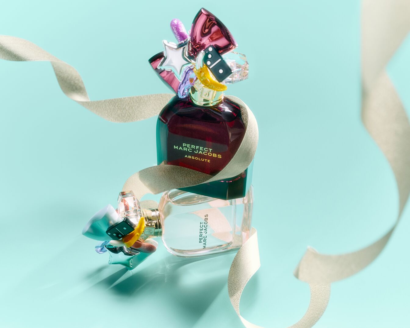 Két üveg Marc Jacobs Perfect parfüm egymásra helyezve, az Absolute sötétvörös, a másik átlátszó, mindkettőjüket szeszélyes charmok díszítik, és egy krémszínű szalag fonja körbe őket világoskék háttér előtt.