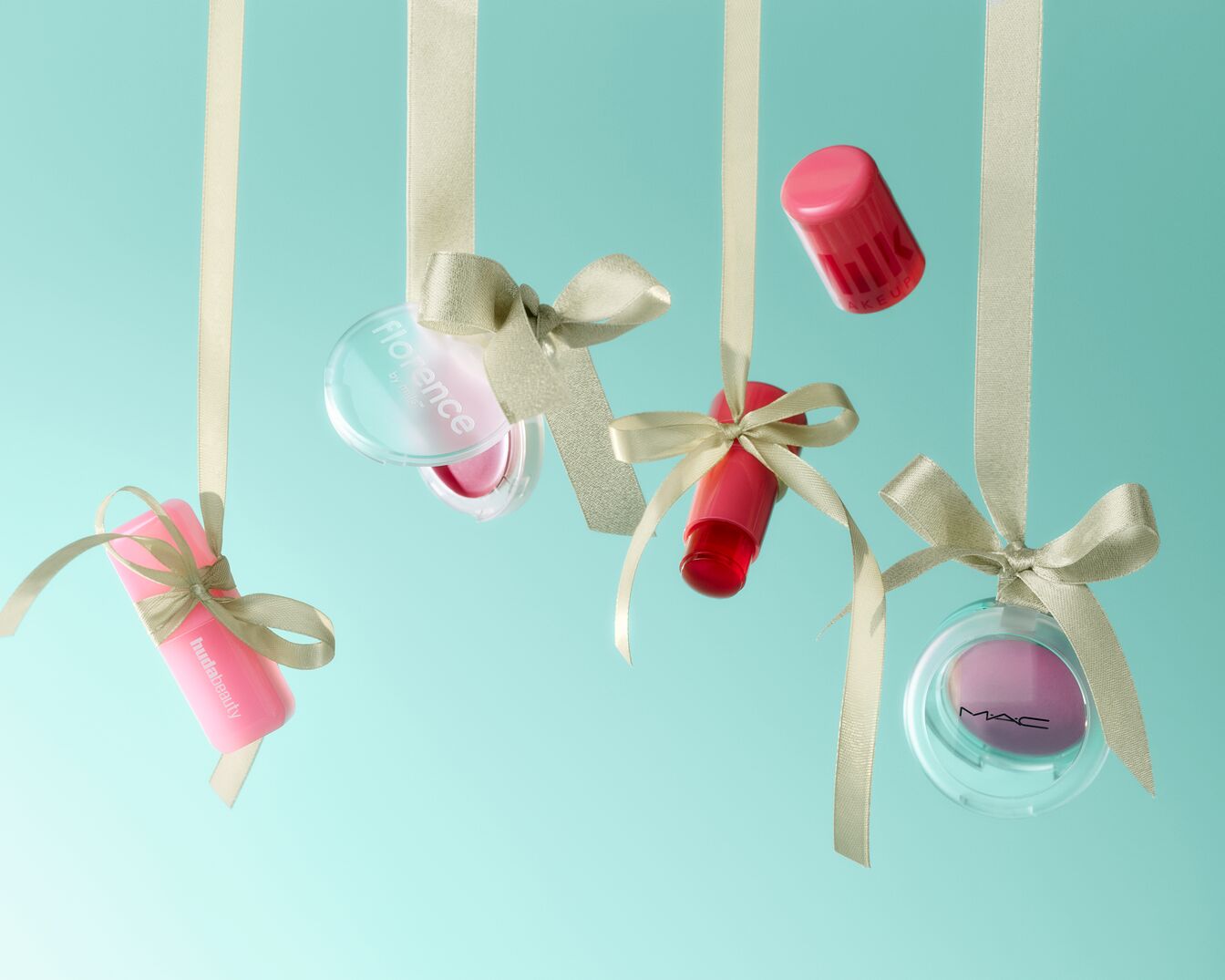 Šventiškai pakabinti kosmetikos gaminiai, įskaitant „Huda Beauty“ ir „Florence by Mills“ produktus, papuošti kreminės spalvos kaspinais ant šviesiai mėlyno fono.