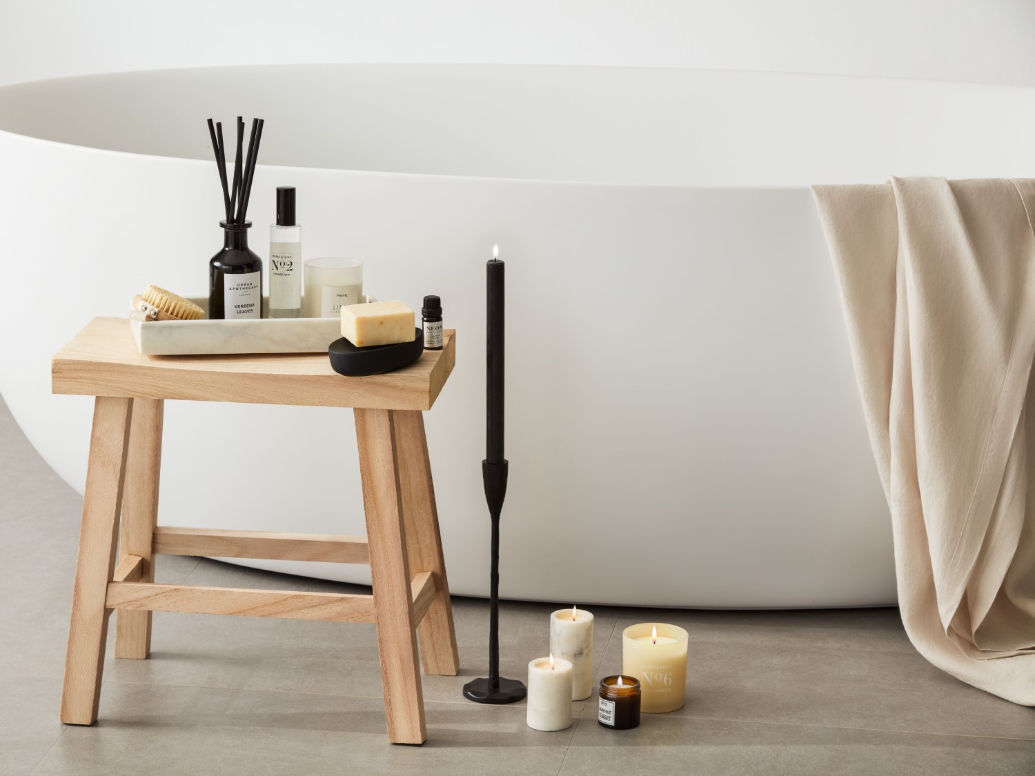 Vonios kambario kompozicija su žvakėmis, balta vonia ir mediniu staliuku, ant kurio padėtas difuzorius, kremas, muilas ir šepetys.