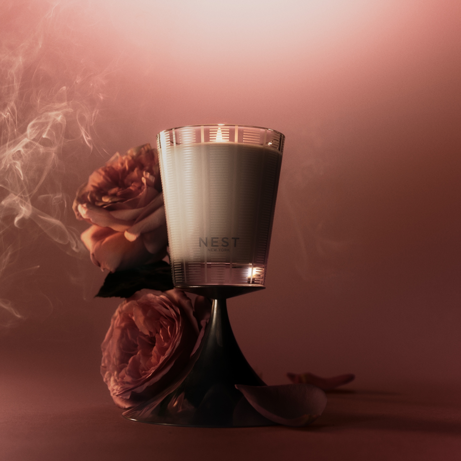 Świeca NEST New York w szklanym, prążkowanym pojemniku, umieszczona na czarnej podstawce, otoczona różami i dymem, tworząca ciepłą, zmysłową atmosferę.
