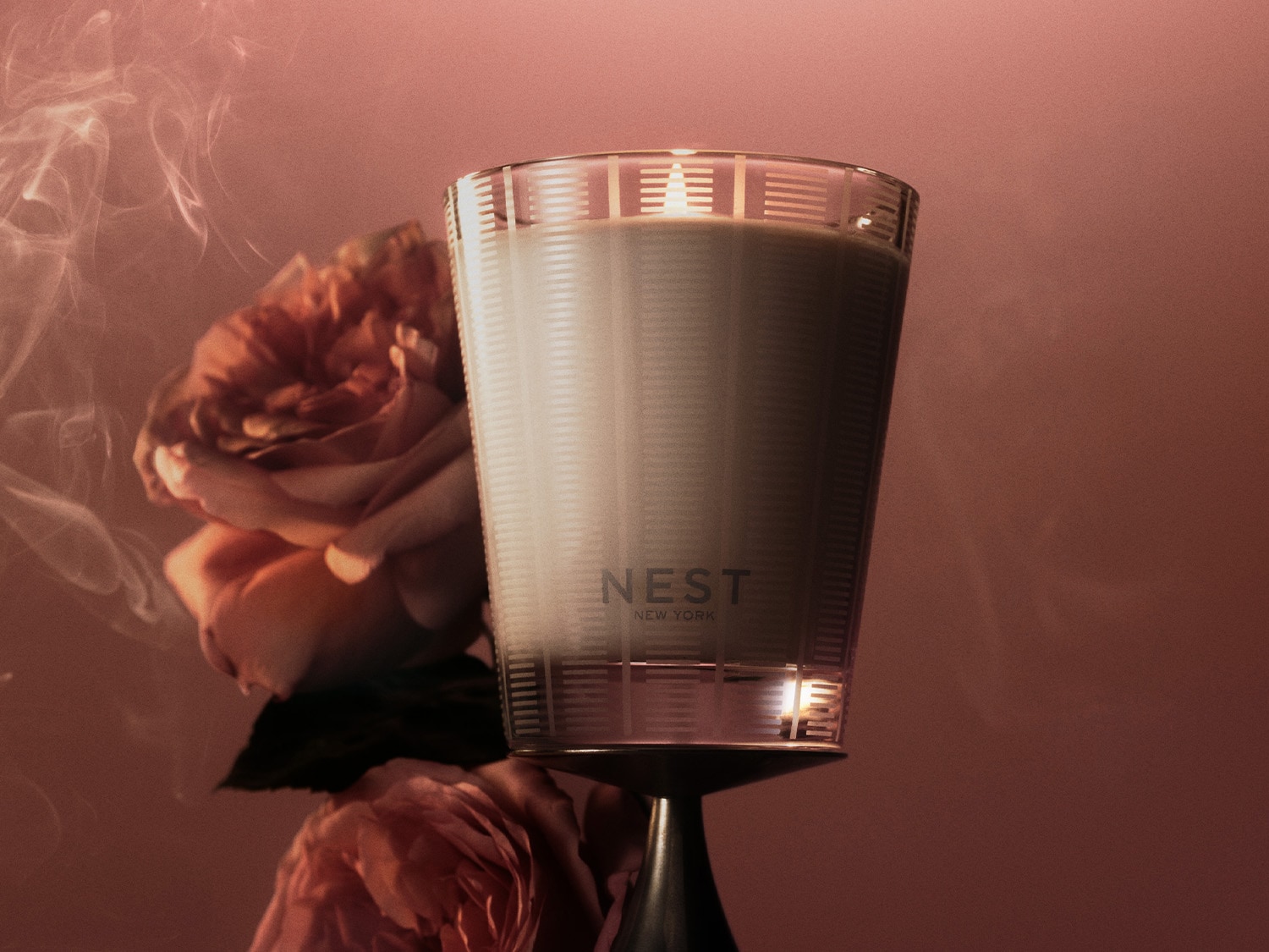 A Nest New York gyertya egy elegáns üvegpohárban, finom rózsaszín virágok és lágy füst kíséretében, meghitt hangulatot teremtve.