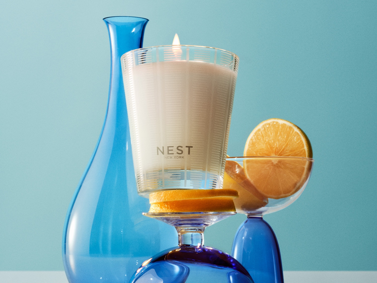 A Nest New York gyertya egy átlátszó üvegben, narancssárga szeletekkel és kék üvegvázával körülvéve, frissítő hangulatot áraszt.