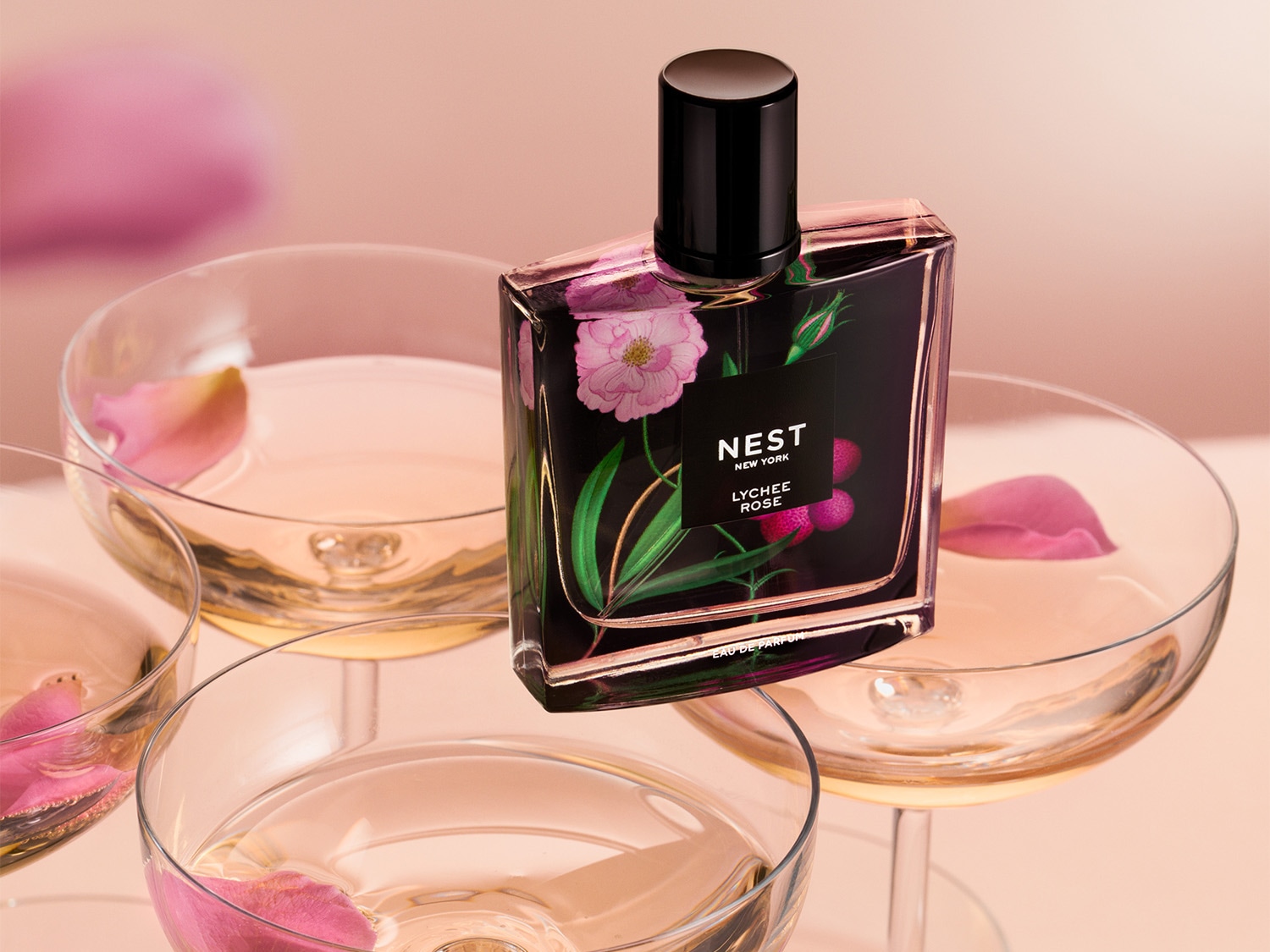 A Nest New York Lychee Rose parfümös üvege virágmintával, rózsaszín szirmokkal teli pezsgőspoharak között.