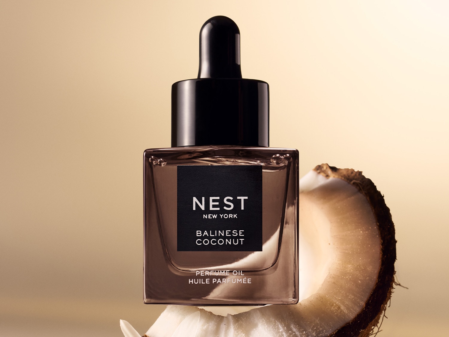 A Nest New York Balinese Coconut parfümolaj egy négyzet alakú, barna üvegben, fekete kupakkal és pipettával, kókuszdió szelet mellett.