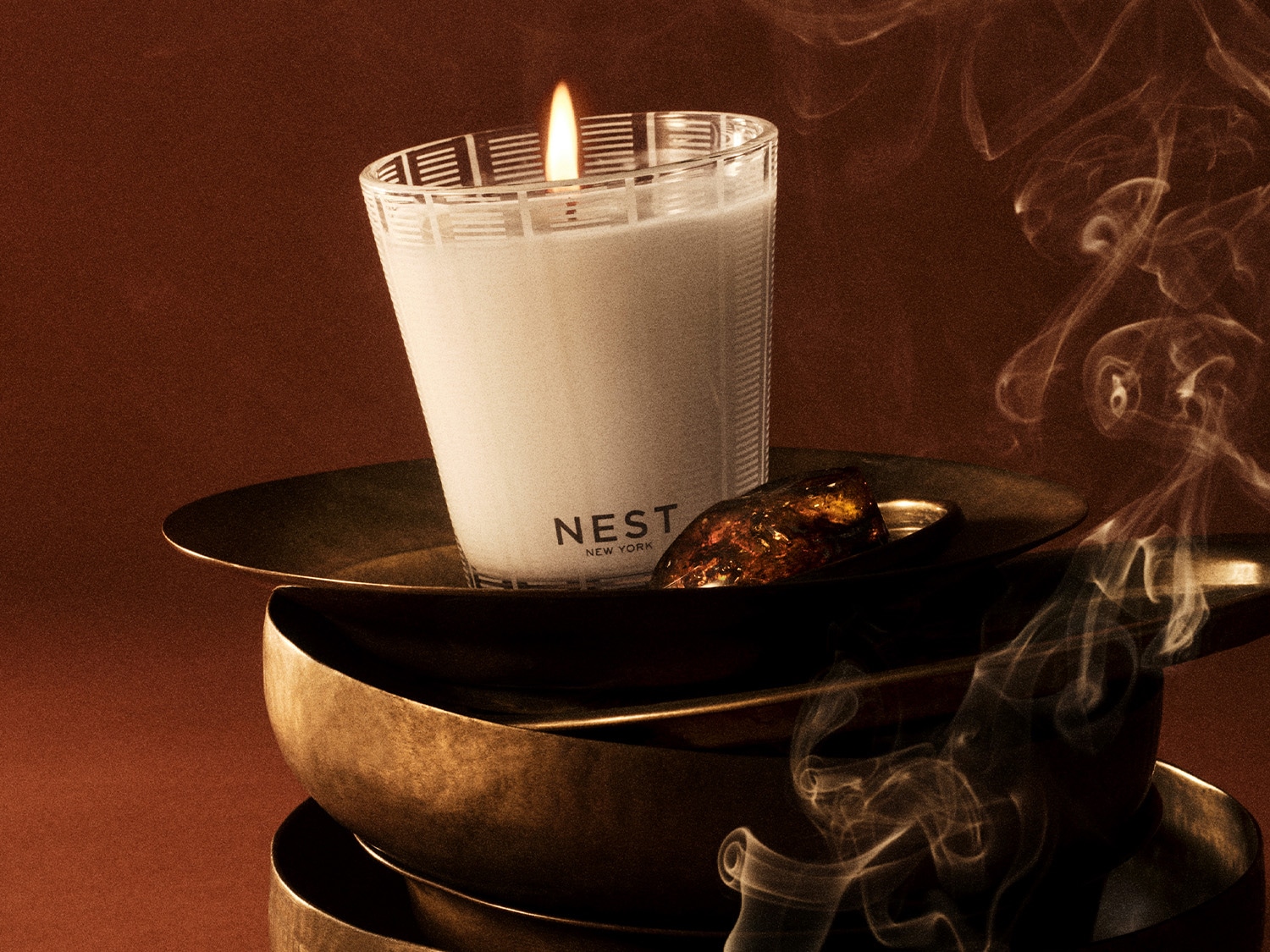 A Nest New York gyertya egy átlátszó üvegben, geometrikus mintákkal, egy arany tálon ül, mellette egy borostyánszínű kődarab, füst gomolyog körülötte, mindez egy réz tálakból álló tornyon.