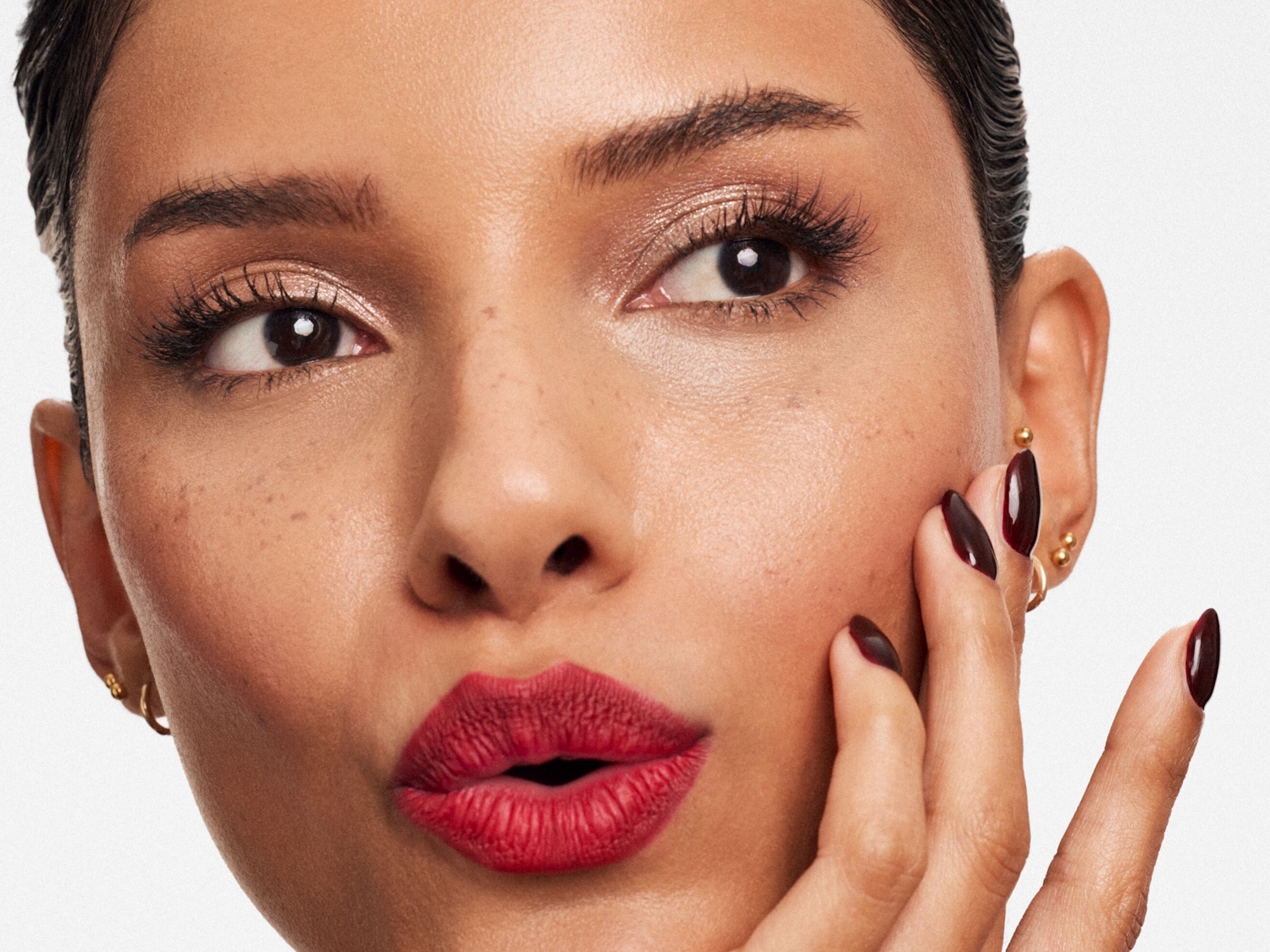 Nahaufnahme eines Gesichtes mit geraden Augenbrauen, goldenem Lidschatten, roten Lippen und dunkelroten Nägeln, die das Gesicht berühren.