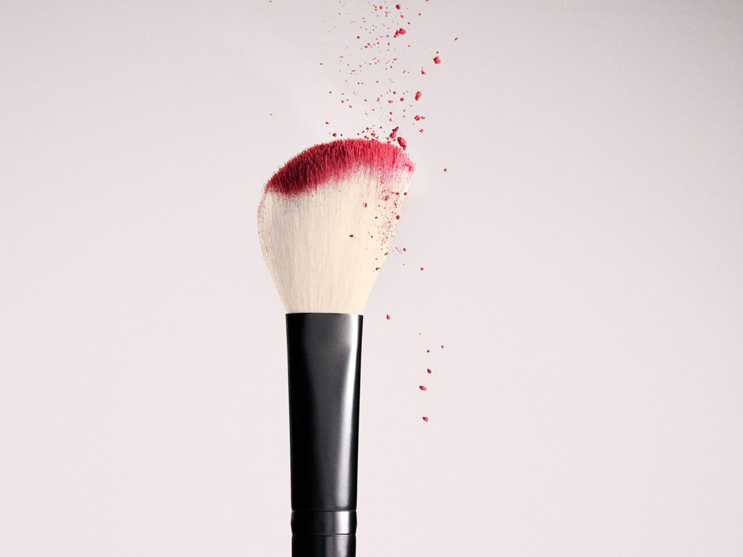 Gros plan d'un pinceau de maquillage avec des poils blancs et un manche noir, dont le bout est recouvert de poudre rose qui s'envole, sur un fond clair.