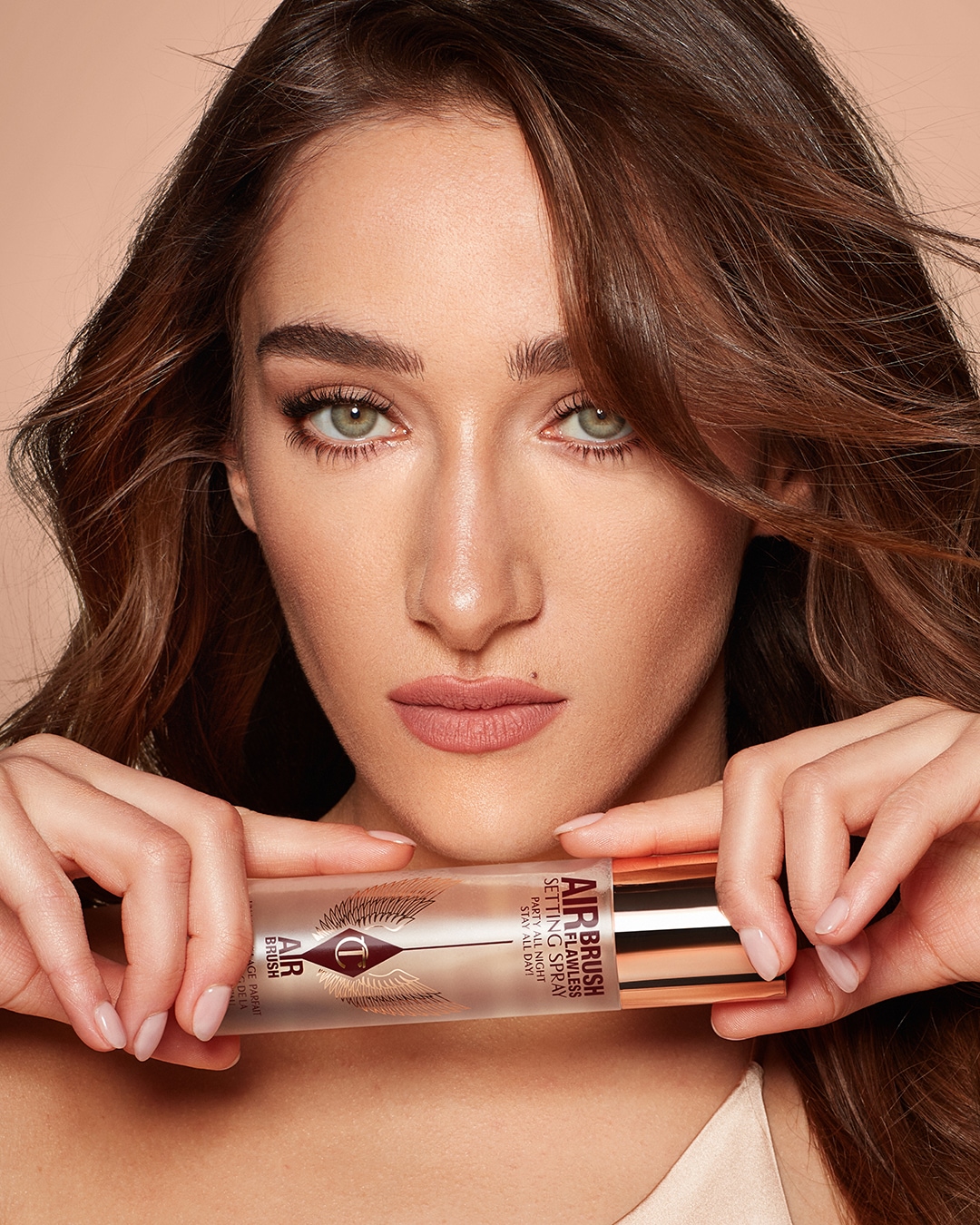 Eine Frau mit braunem, gewelltem Haar und grünen Augen hält Charlotte Tilbury Airbrush Flawless Setting Spray in ihren Händen, das verspricht, das Make-up die ganze Nacht haltbar zu machen.
