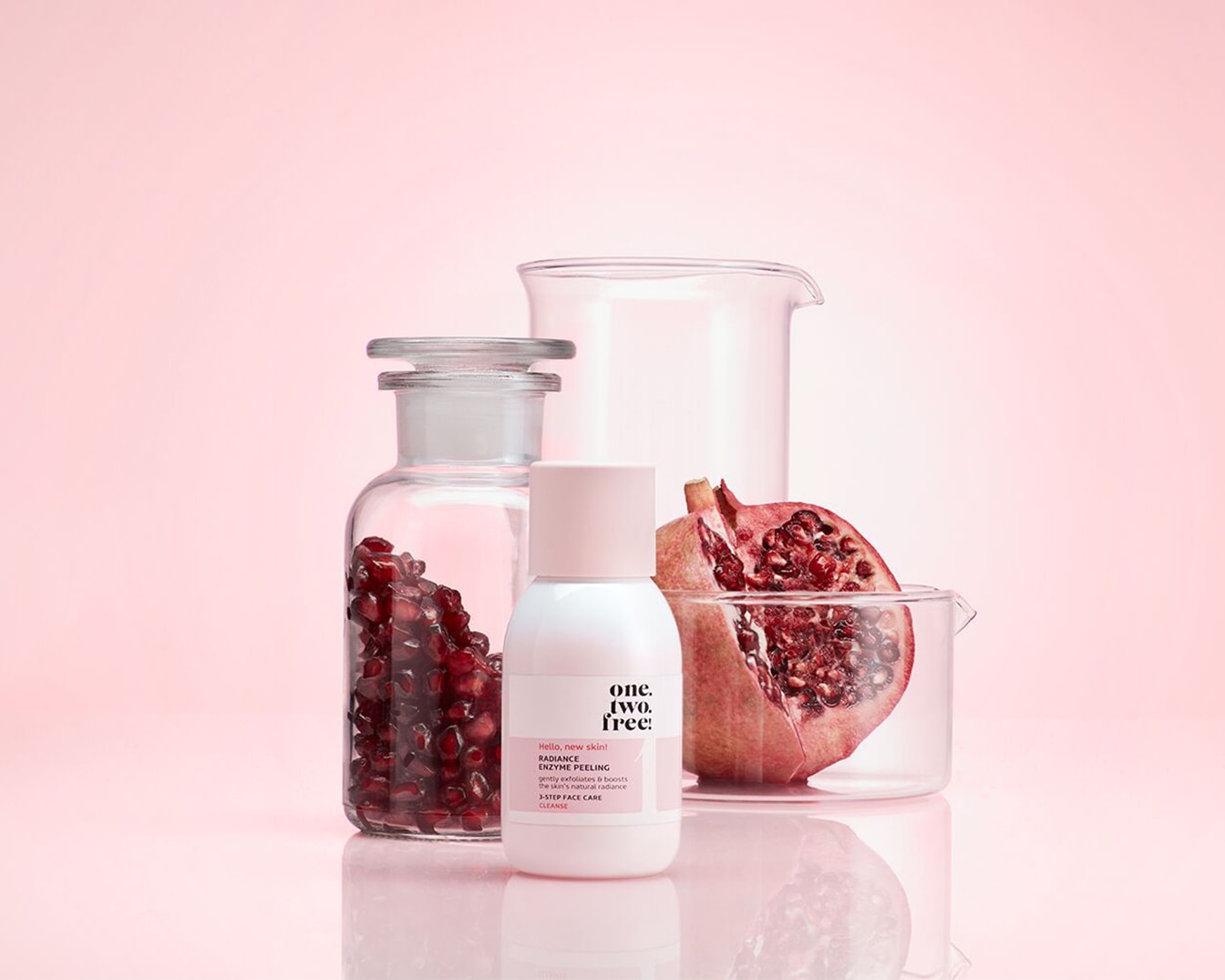 Auf einem rosa Hintergrund befinden sich ein weißer Spender mit rosa Deckel, ein Glas mit Granatapfelkernen, ein halber Granatapfel und zwei Messbecher.