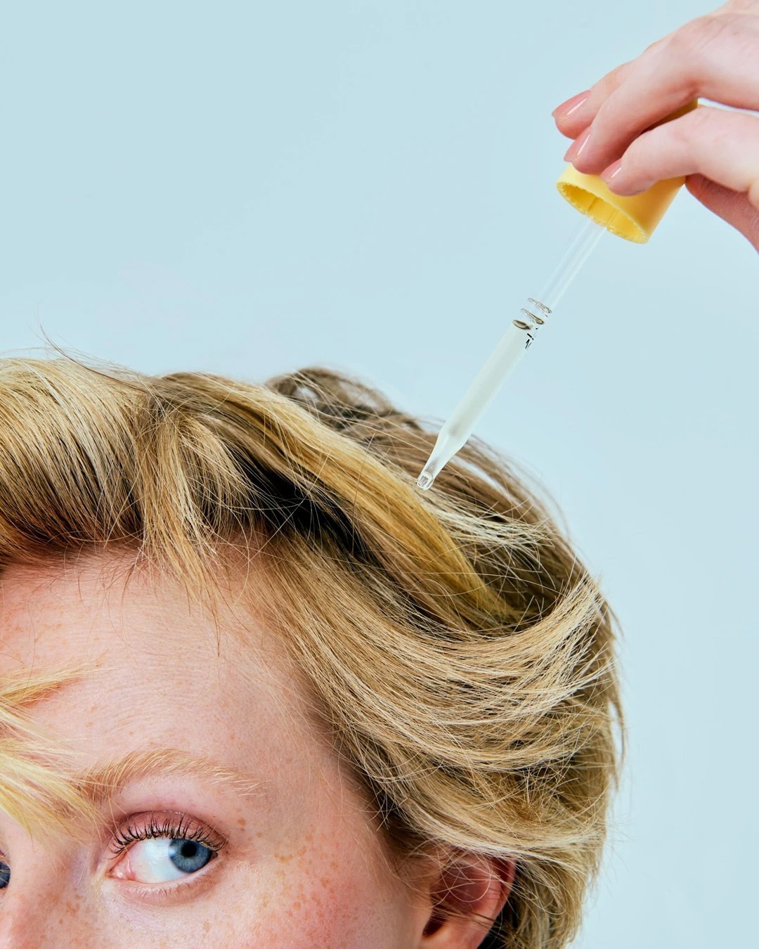 Gros plan sur une personne aux cheveux blonds recevant un traitement à l'huile capillaire à l'aide d'une pipette jaune, pour des cheveux nourris et éclatants.