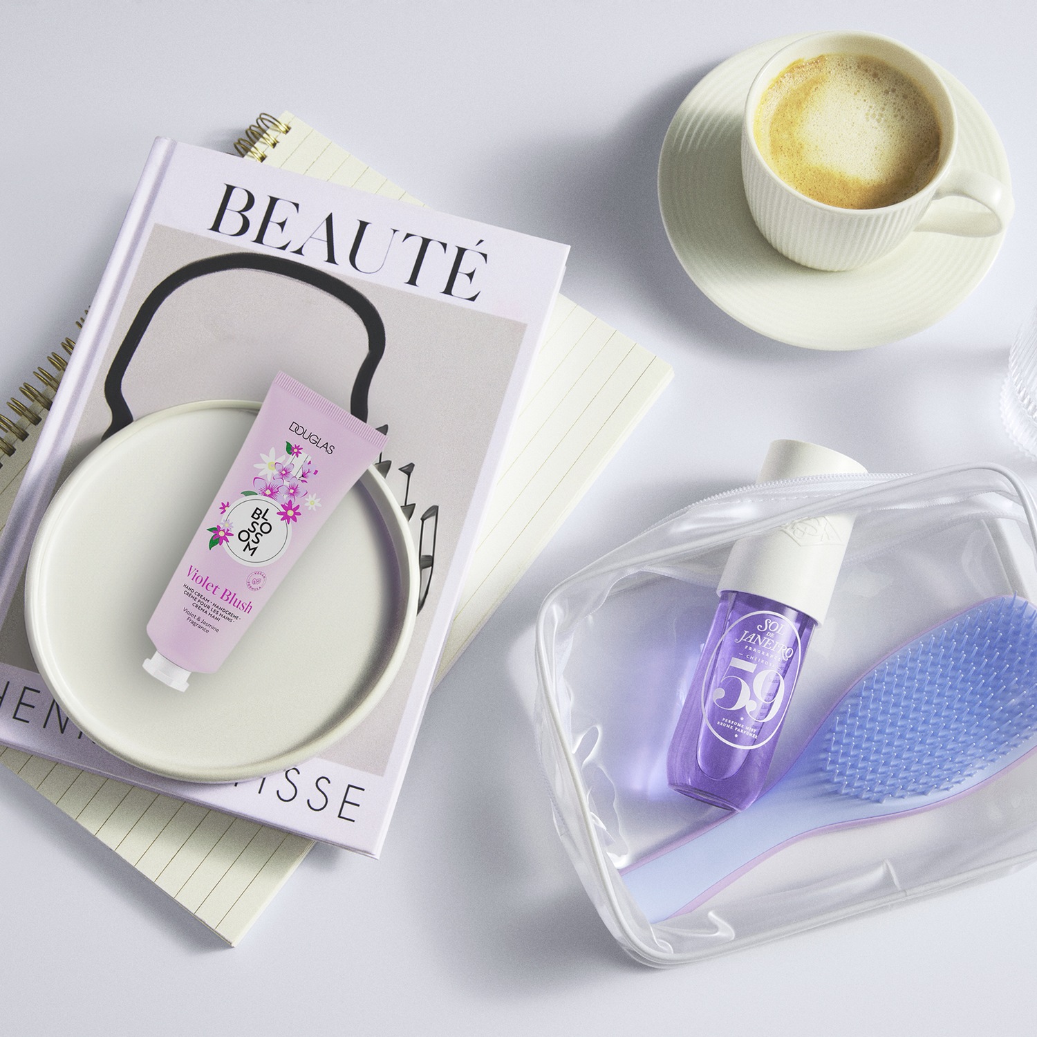 Een platliggende scène met een Douglas Violet Blush handcrème op een boek, een kop koffie en een transparant tasje met een Sol de Janeiro parfum en een haarborstel.