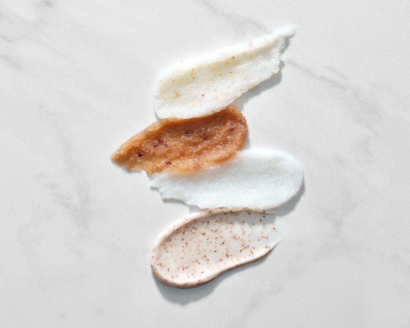 Keturių skirtingų spalvų ir tekstūrų šveitiklių tepinėliai ant balto marmurinio paviršiaus.