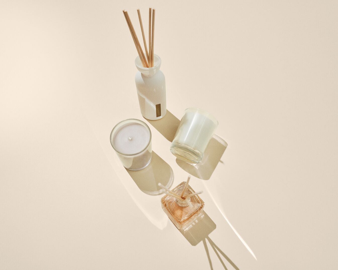 Nuotraukoje matomi kvapų difuzoriai su lazdelėmis ir žvakės stikliniuose indeliuose, išdėstyti ant šviesaus paviršiaus, sukuriančio švelnius šešėlius.