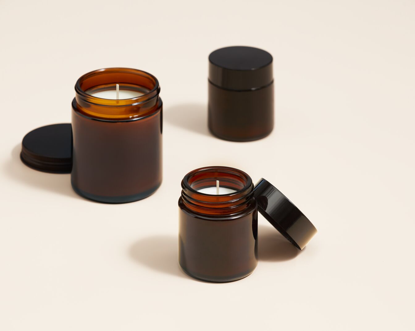 Trys rudos žvakės stikliniuose indeliuose su juodais dangteliais ant smėlio spalvos paviršiaus sukuria jaukią ir raminančią atmosferą.