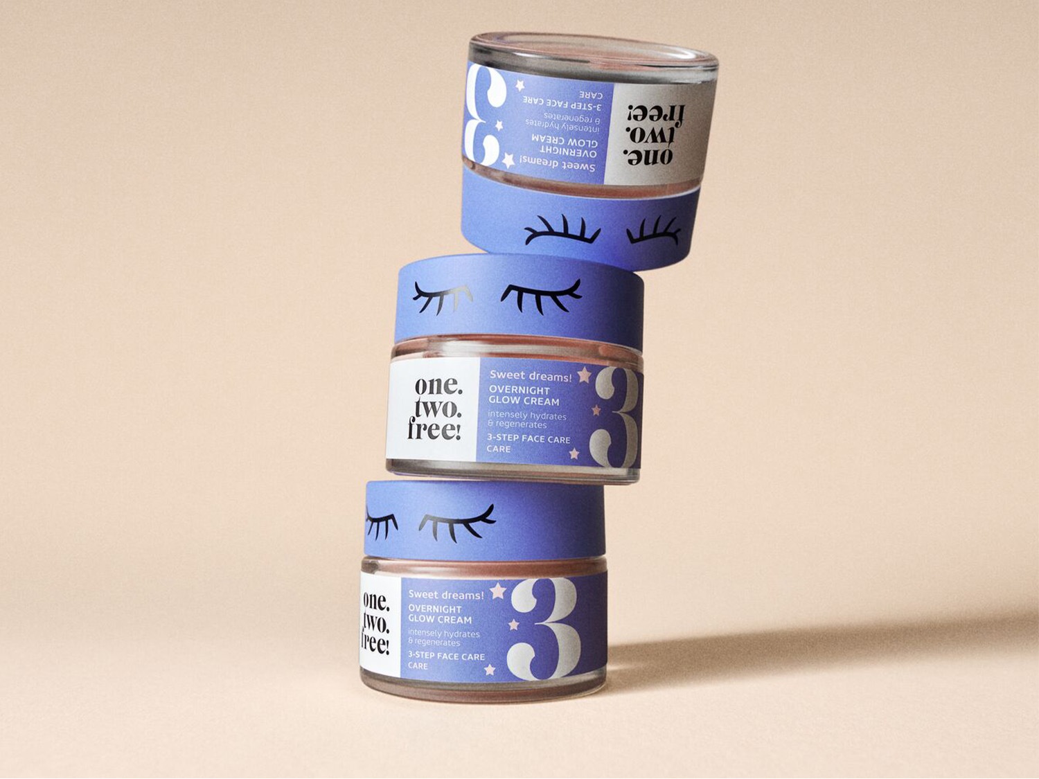 Voici une pile de trois pots de crème de nuit One.Two.Free! avec des couvercles bleu lavande décorés de cils endormis, promettant d'hydrater et de régénérer la peau pour des rêves doux.