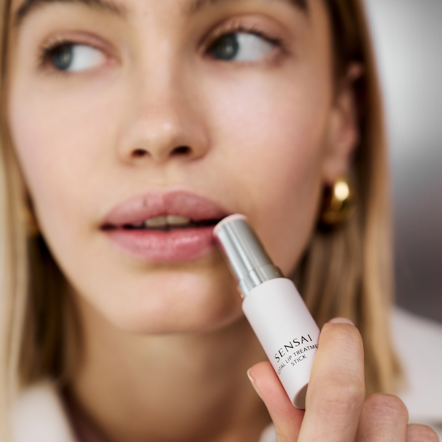 Eine Frau mit blonden Haaren trägt einen Lippenpflegestift von Sensai auf, um ihre Lippen zu pflegen.