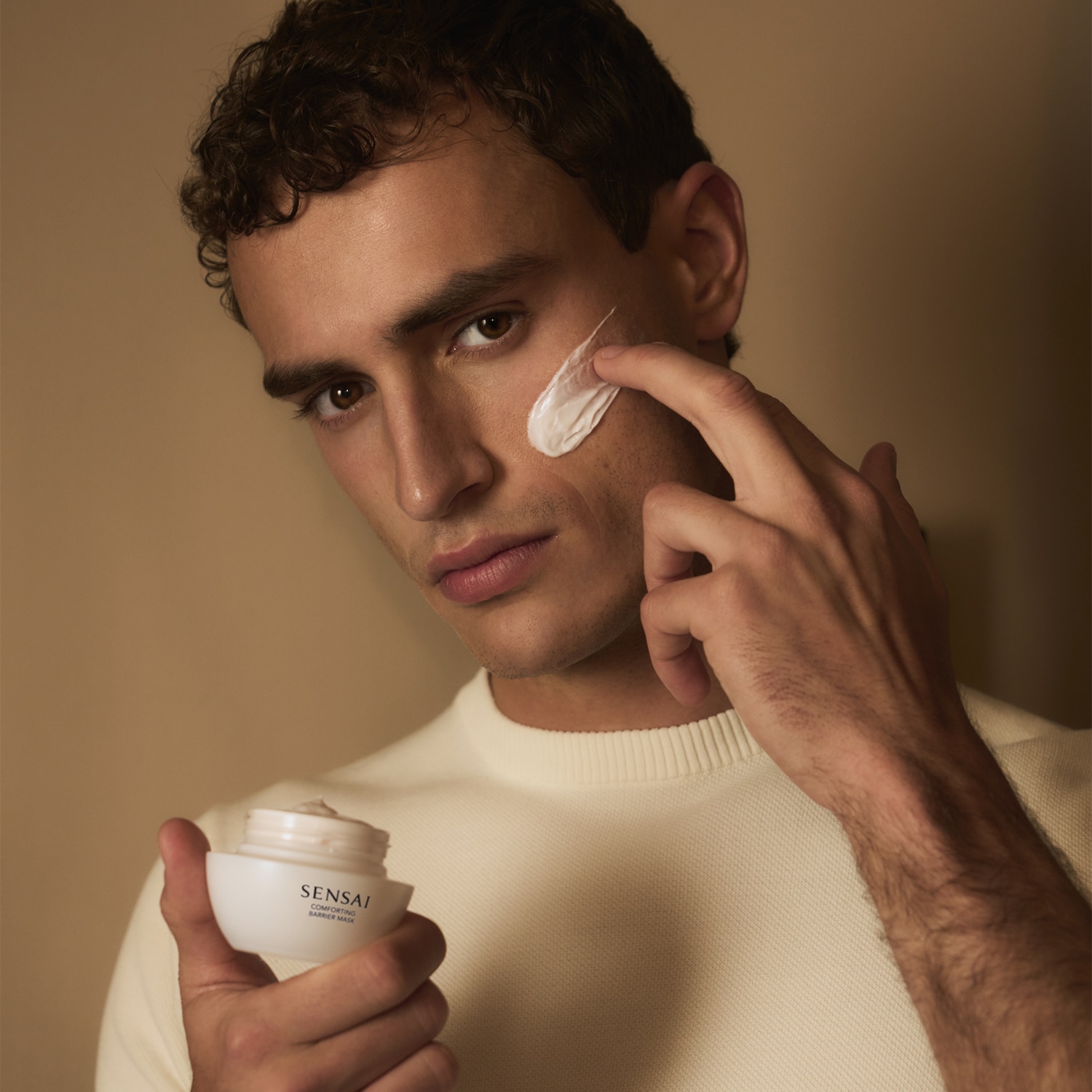 Ein Mann mit lockigem Haar trägt eine weiße Creme aus einem weißen Tiegel auf seine Wange auf, um seine Haut zu pflegen.