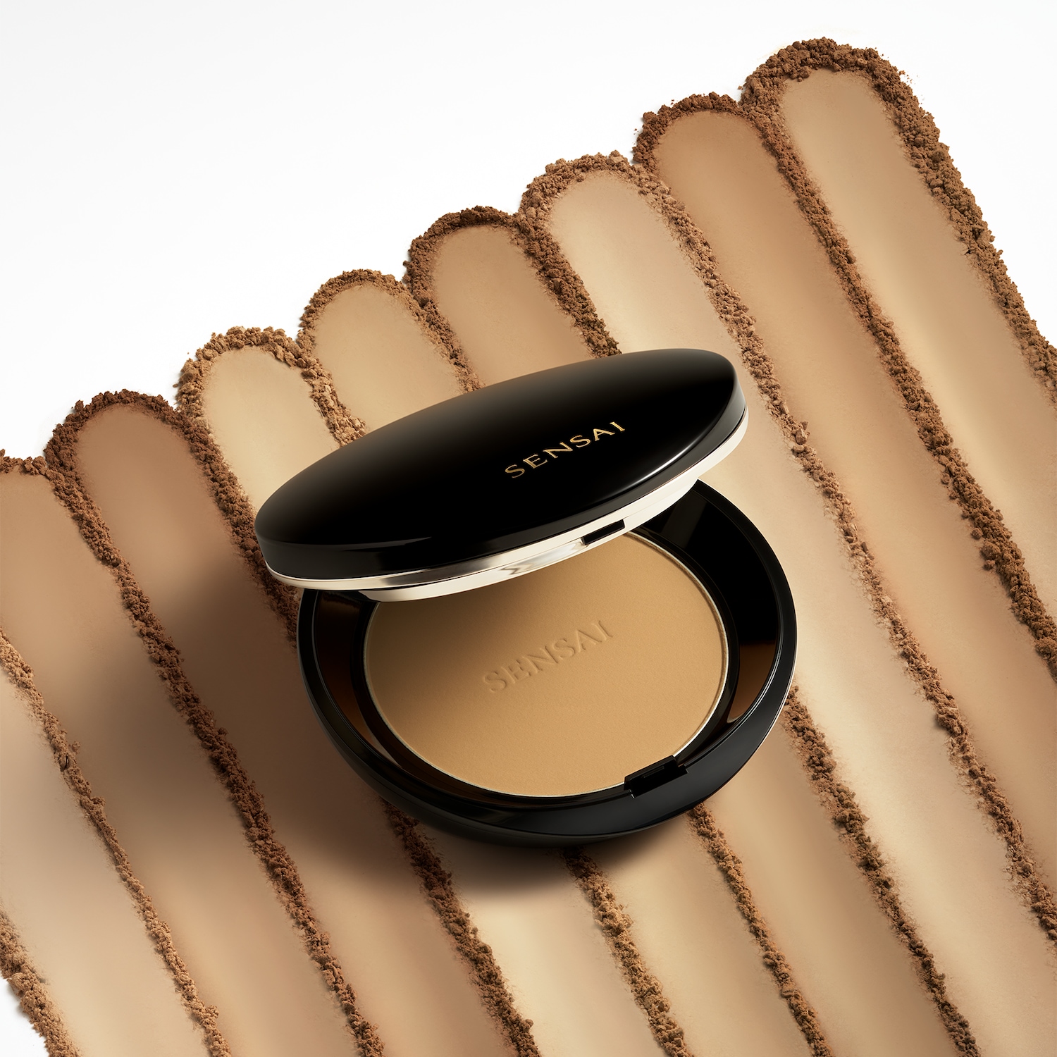 Das Kompaktpuder von Sensai in einer schwarzen Puderdose mit geöffnetem Deckel, umgeben von Puderstreifen in verschiedenen Brauntönen.