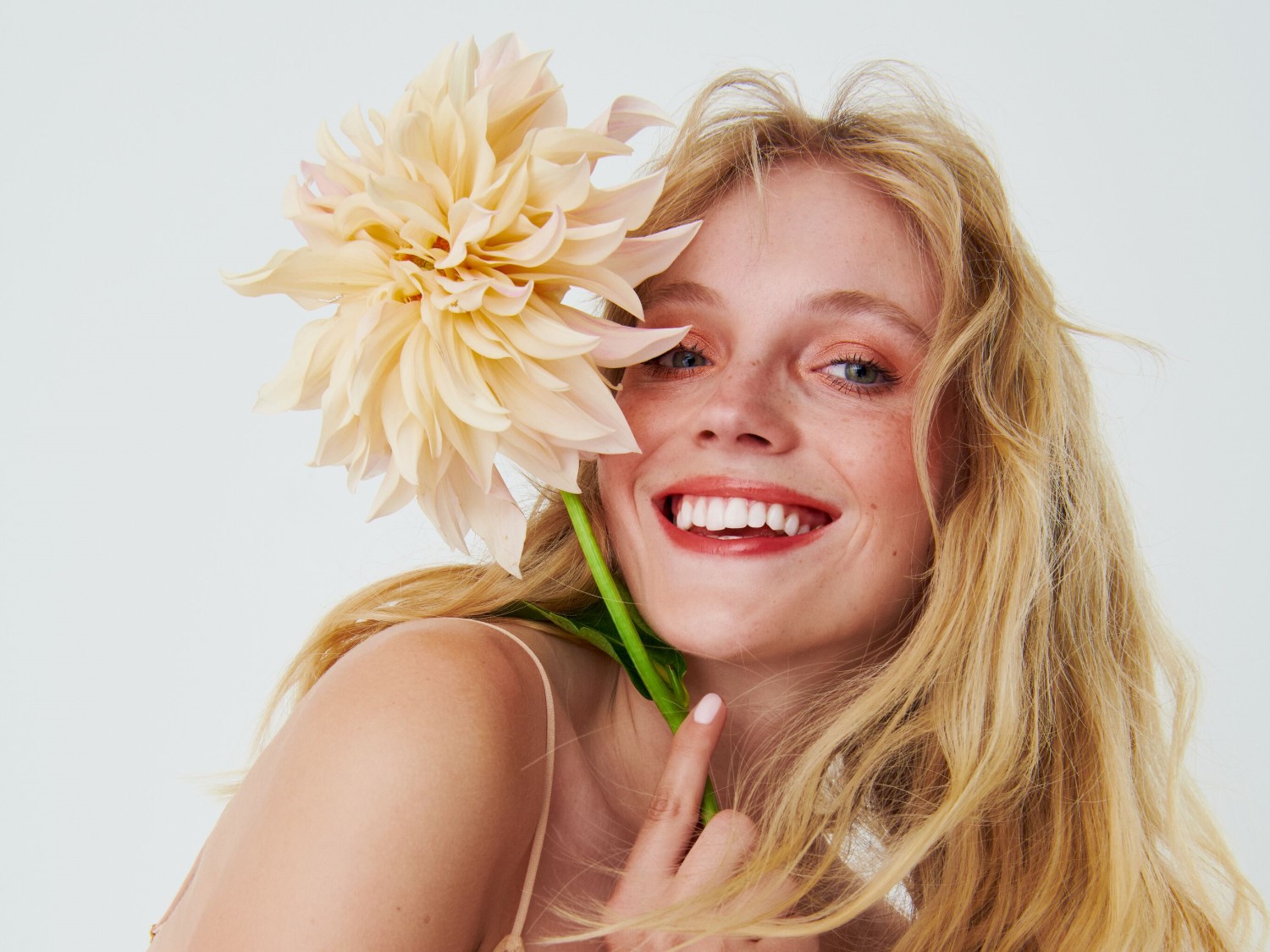 Eine strahlende Frau mit blonden, gewellten Haaren lächelt und hält eine große, hellgelbe Blume an ihr Gesicht.