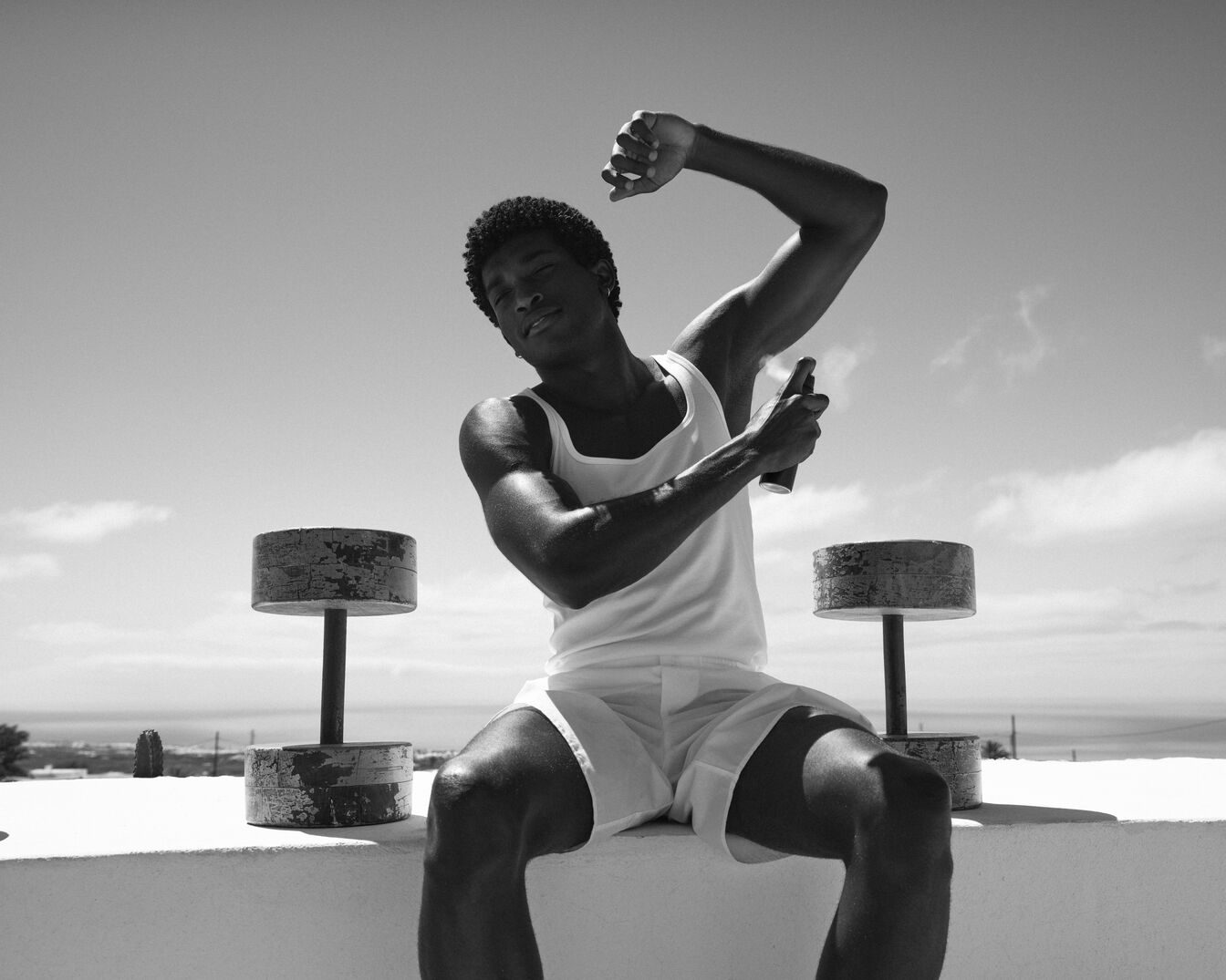 Juodai balta nuotrauka vyro, sėdinčio ant sienos su svarmenimis šalia, purškiančio dezodorantą ant pažasties.