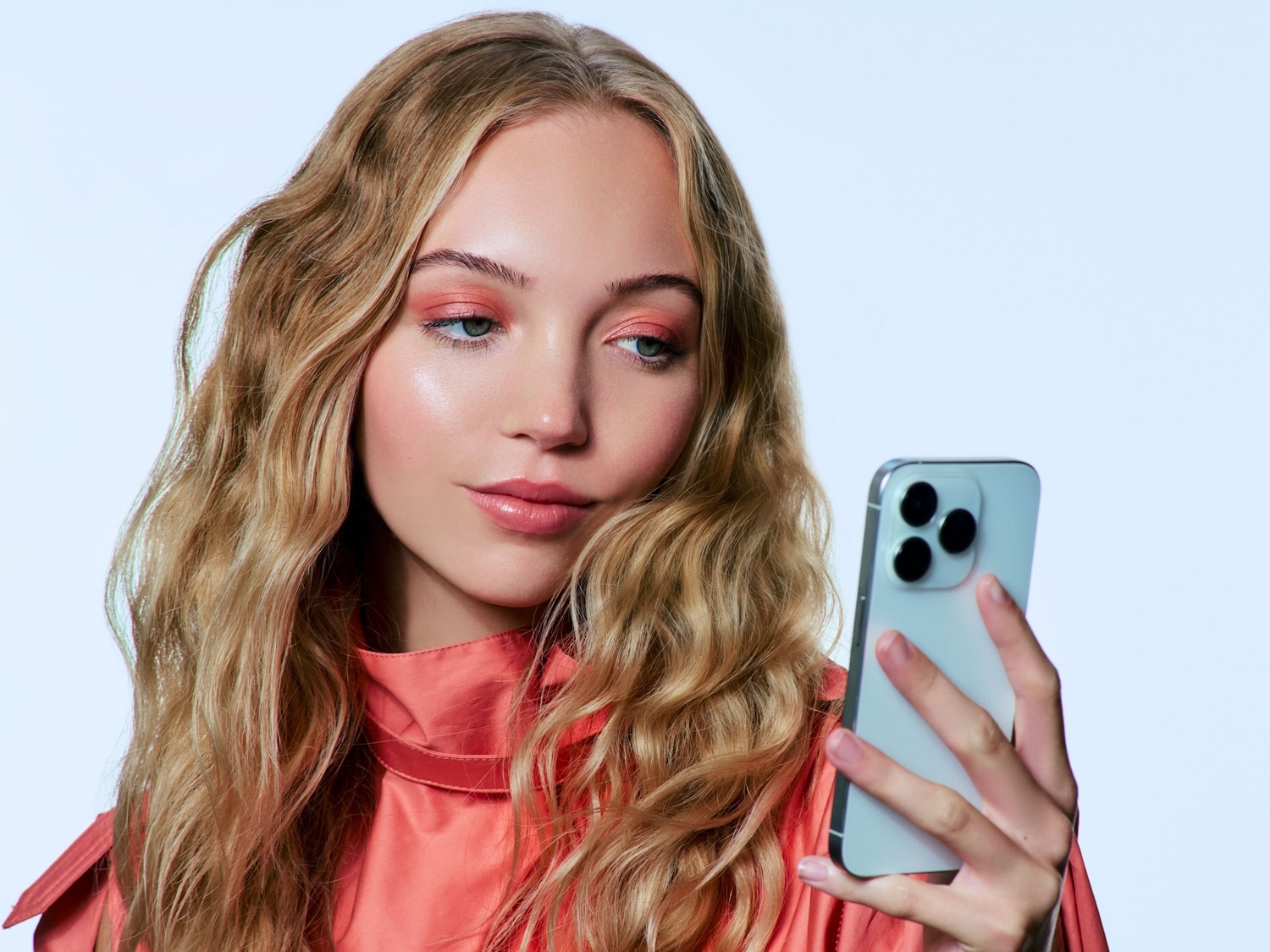Eine blonde Frau mit gewelltem Haar und einem korallenroten Oberteil hält ein Smartphone in der Hand und hat ein peachiges Make-up.