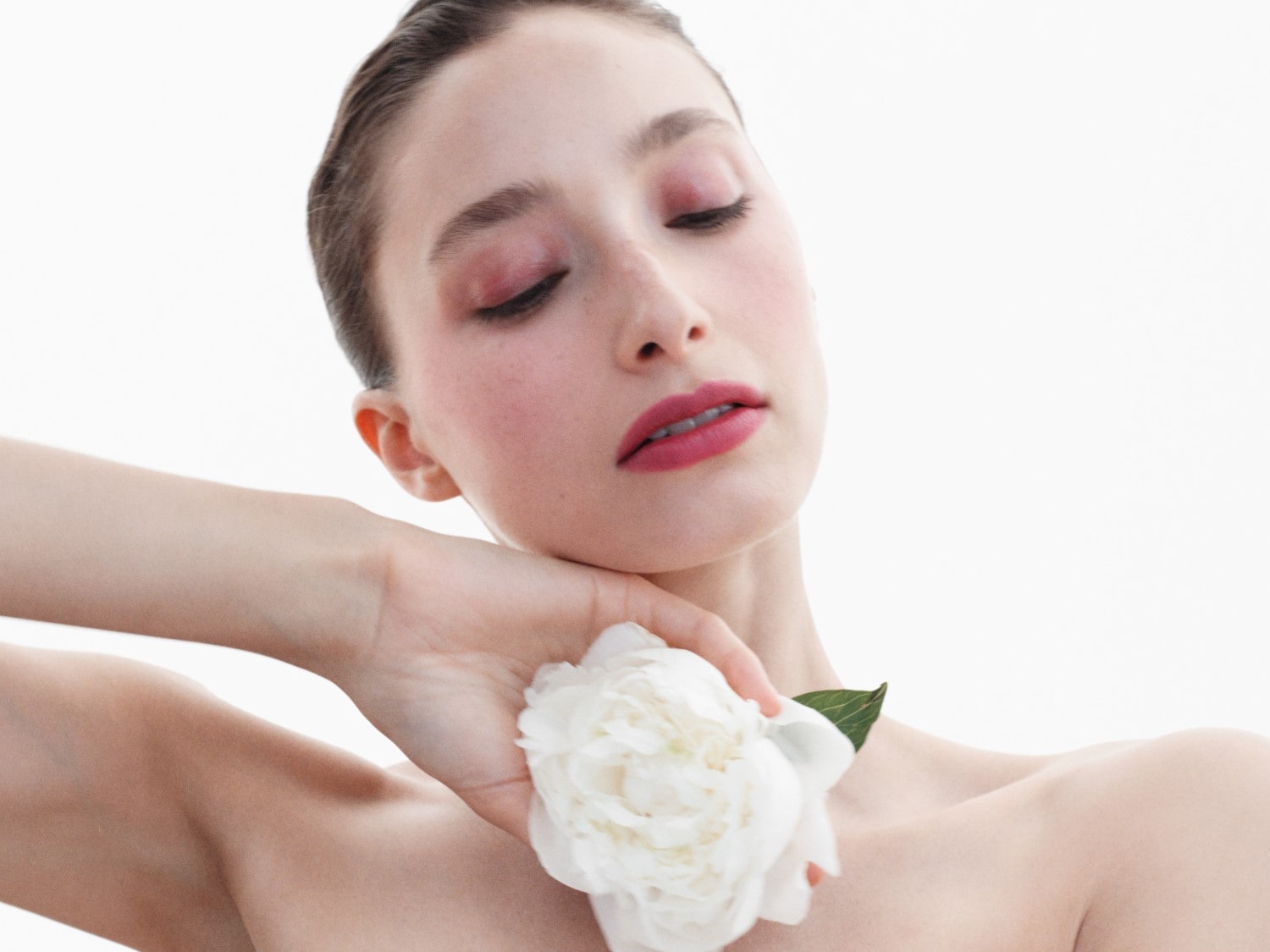 Eine Frau mit heller Haut hält eine weiße Blume an ihrem Hals, ihr Make-up ist in sanften Rosatönen gehalten.