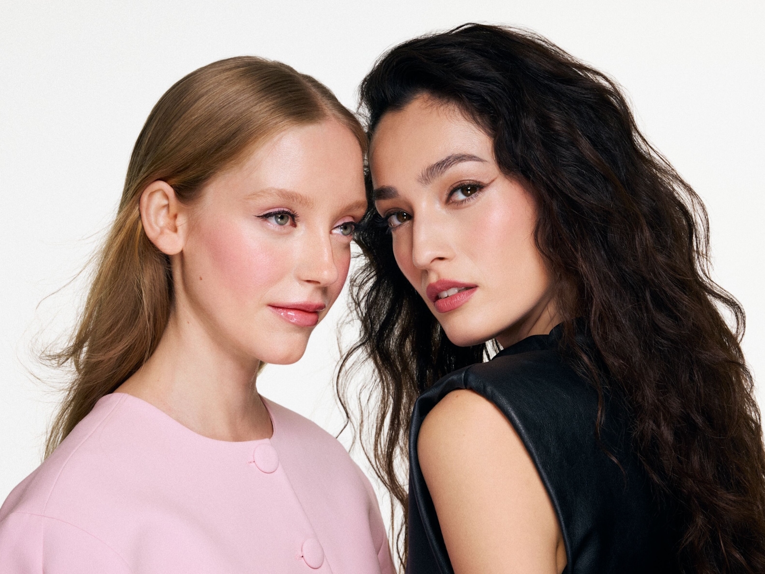 Zwei Frauen mit zarter, rosafarbener Schminke und glänzenden Lippen, wobei die eine einen rosa Blazer und die andere ein schwarzes Ledertop trägt.