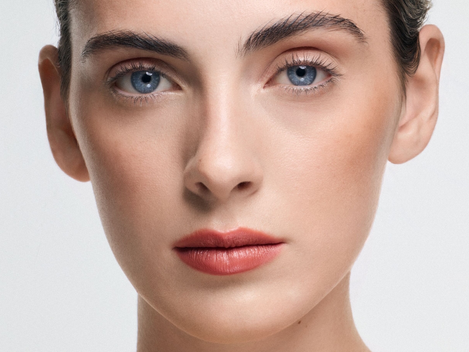 Nahaufnahme eines Frauengesichts mit hellblauen Augen, vollen Lippen mit korallfarbenem Lippenstift und gepflegten Augenbrauen.