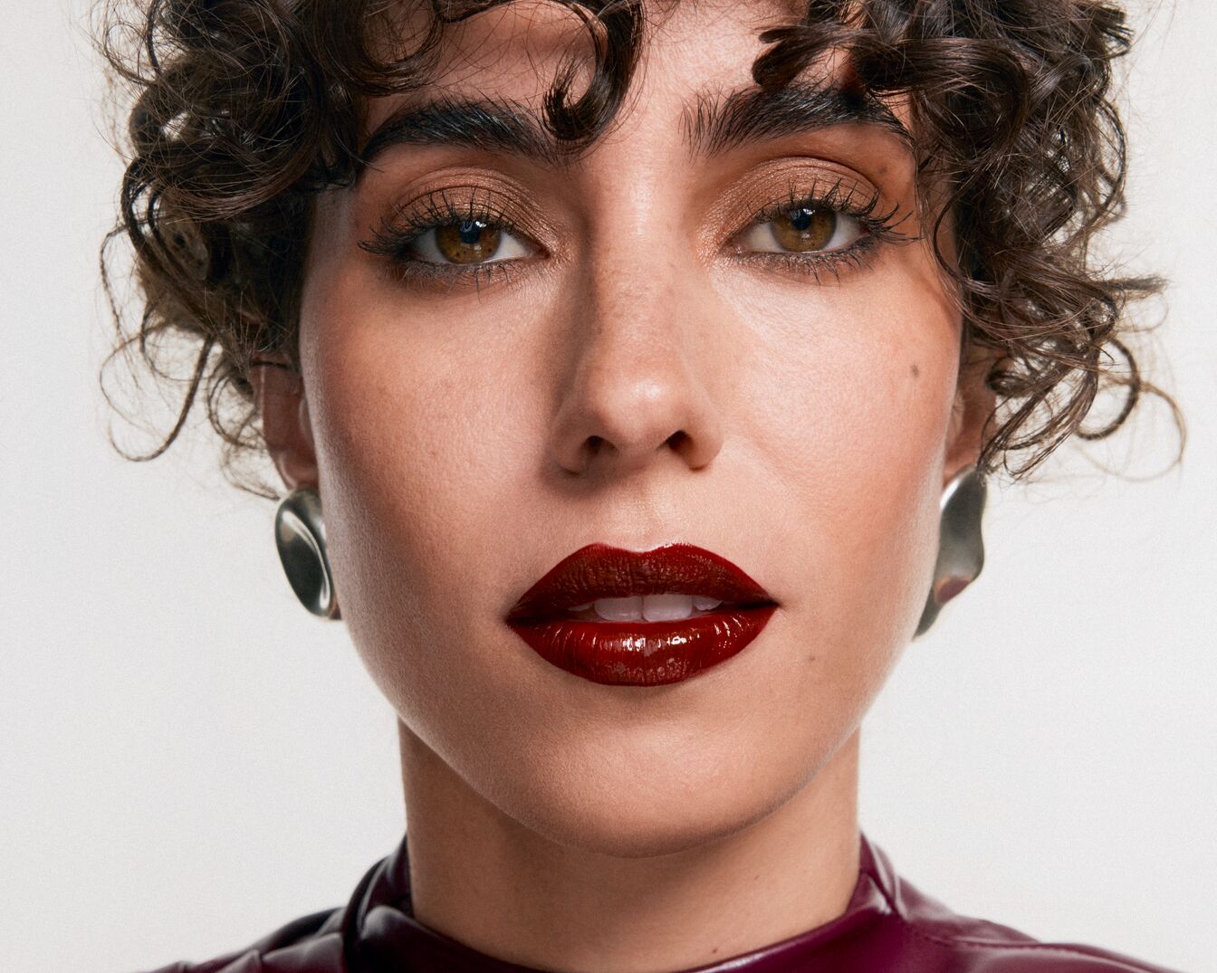 Nahaufnahme einer Frau mit lockigem, braunem Haar, braunen Augen und glänzendem, dunkelrotem Lippenstift, die silberne Ohrringe und ein weinrotes Oberteil trägt.