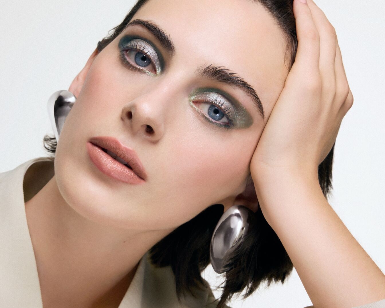 Nahaufnahme einer Frau mit silbernem und grünem Lidschatten, nudefarbenem Lippenstift und großen silbernen Ohrringen, die einen modernen und eleganten Look erzeugt.