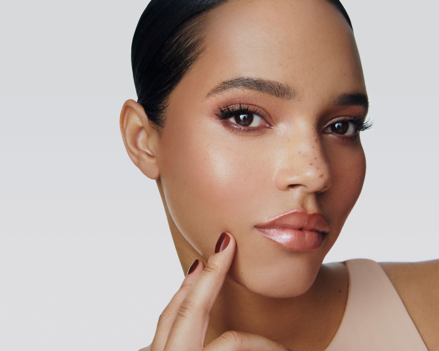 Nahaufnahme einer Frau mit glänzenden Lippen, braunen Augen und braunem Nagellack, die ihre Wange berührt, um ein strahlendes Make-up zu präsentieren.