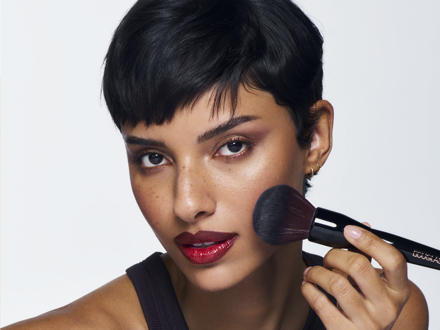 Nahaufnahme einer Frau mit kurzem, dunklem Haar, die mit einem Douglas-Pinsel Rouge auf die Wangen aufträgt, um einen strahlenden Teint zu erzielen.