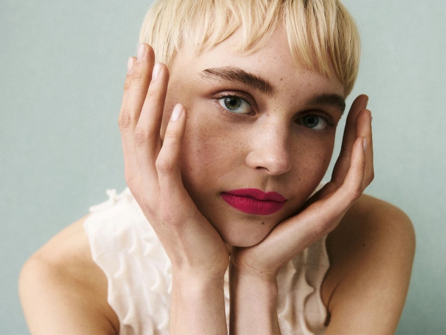 Nahaufnahme einer Frau mit blonden, kurzen Haaren, die ihre Hände an ihr Gesicht legt und einen leuchtend pinkfarbenen Lippenstift trägt.