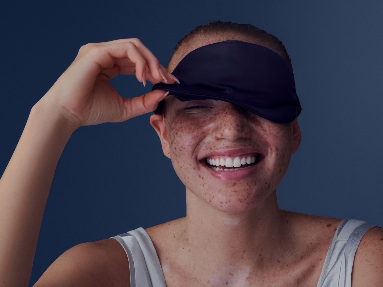 Eine lächelnde Frau mit Sommersprossen hebt eine dunkelblaue Schlafmaske von ihren Augen.