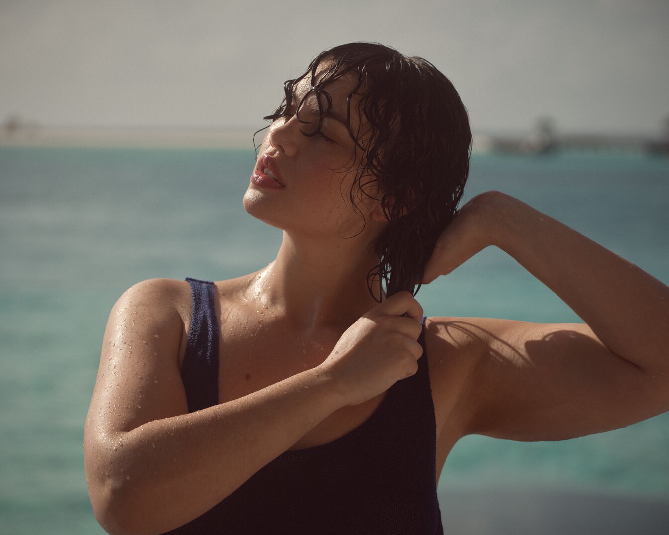 Eine Frau mit nassen, dunklen Haaren und einem marineblauen Badeanzug steht vor dem Meer und genießt die Sonne.