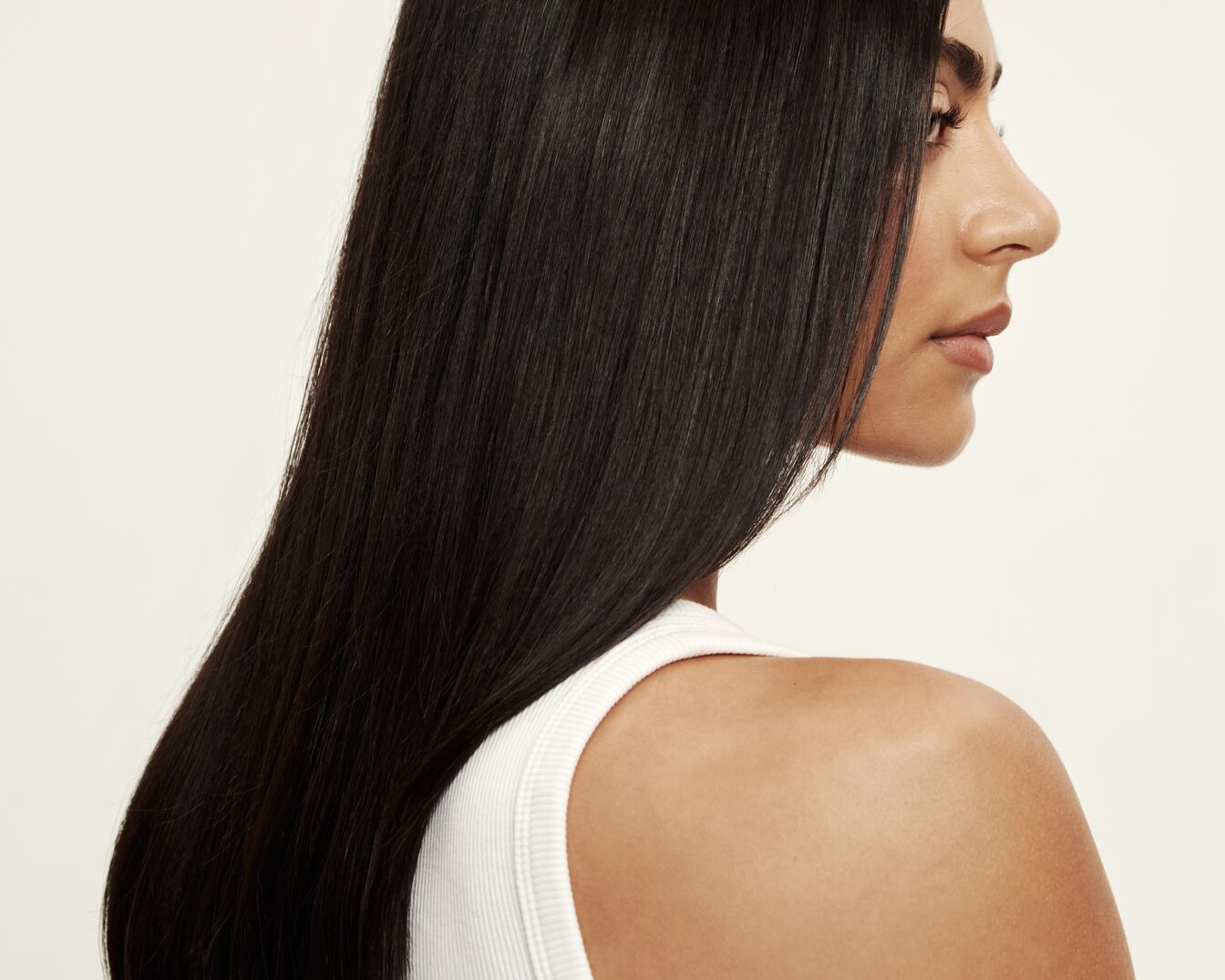 Profil einer Frau mit langem, glänzendem, dunklem Haar und einem weißen Tanktop vor einem hellen Hintergrund.