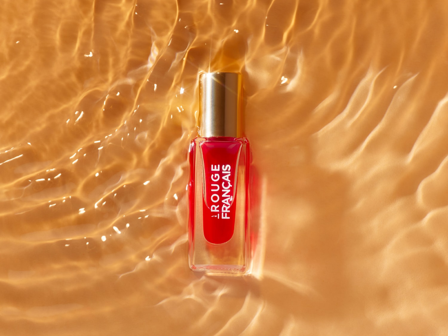 Découvrez le vernis à ongles Rouge Français, un rouge éclatant dans un flacon transparent avec un bouchon doré, flottant sur une surface dorée ondulée.