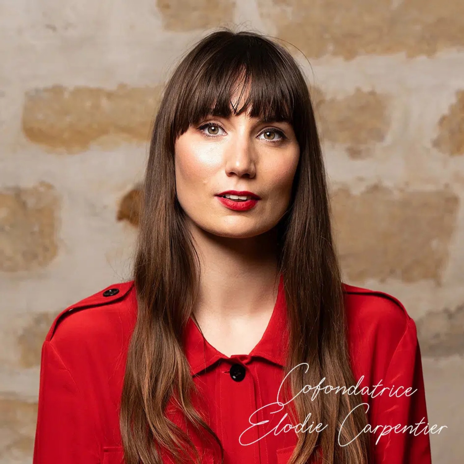 Portrait d'Élodie Carpentier, cofondatrice, portant une chemise rouge et un rouge à lèvres rouge vif, devant un mur en pierre.