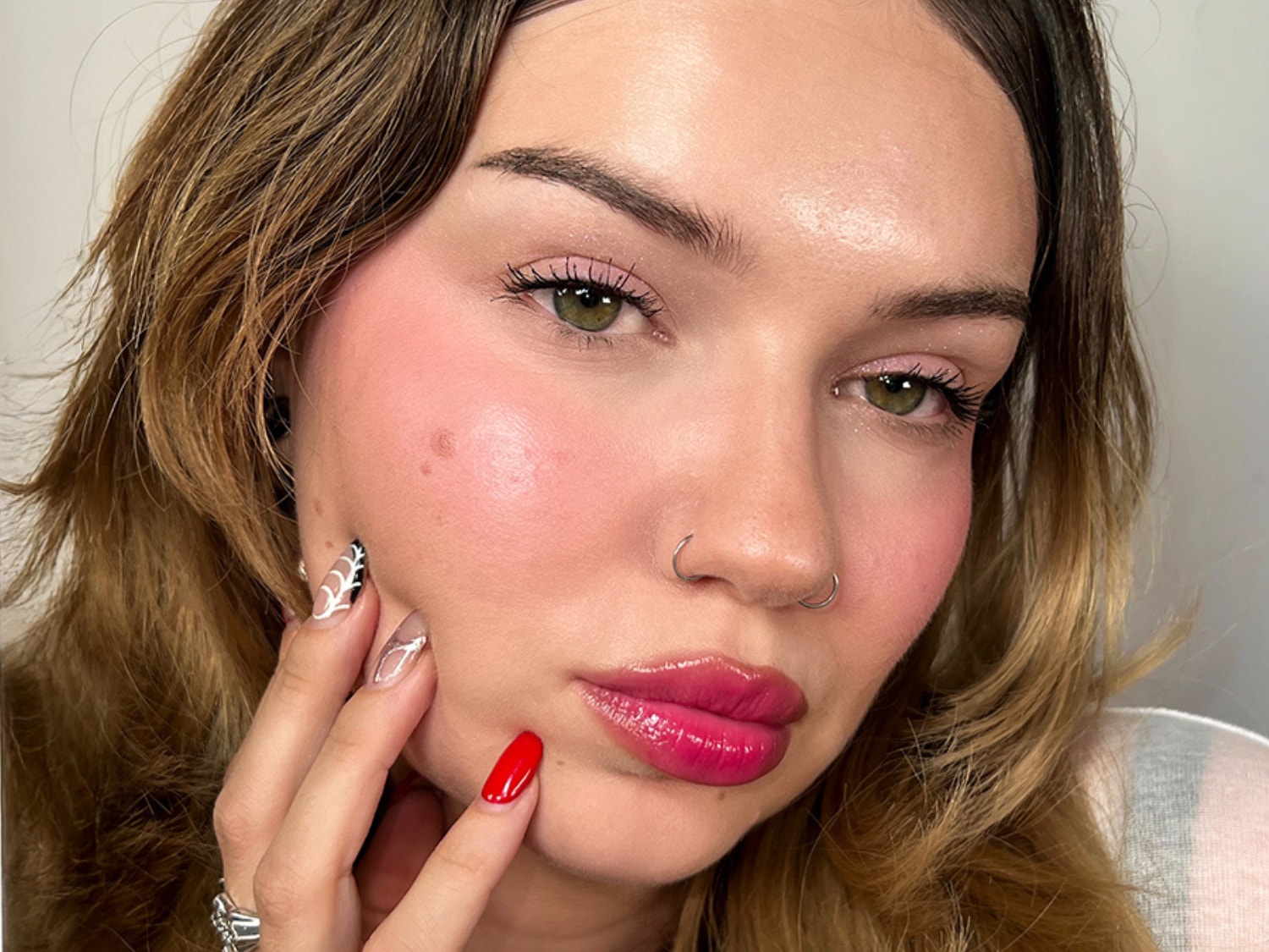 Een close-up van een vrouw met een frisse make-uplook, roze blosjes op de wangen en glanzende roze lippen, die haar gezicht zachtjes aanraakt met haar hand.
