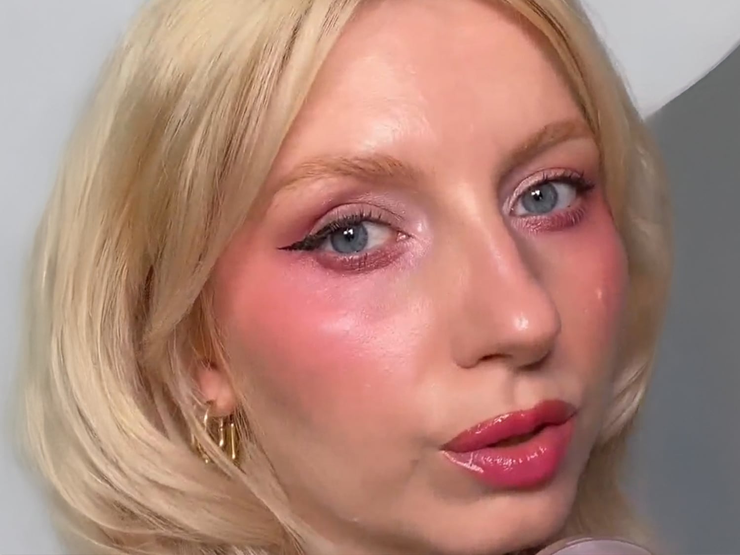 Een close-up van een vrouw met blonde haren, blauwe ogen en roze make-up op haar ogen, wangen en lippen, gecombineerd met een zwarte eyeliner.