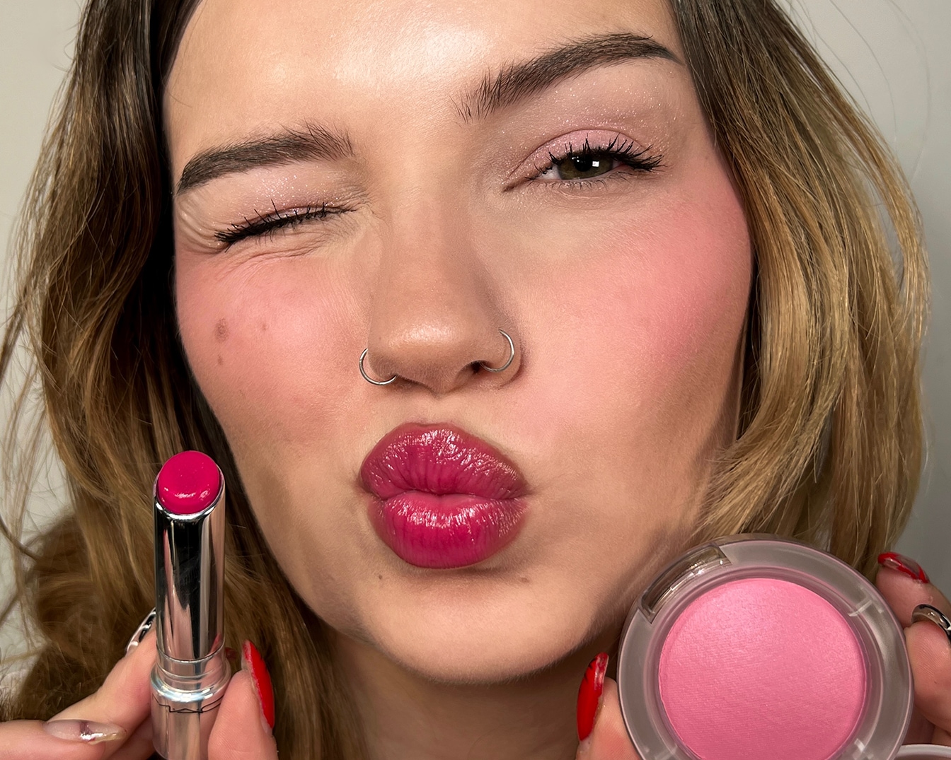 Een vrouw met een knipoog, roze lippenstift en blosjes, houdt een zilveren lippenstift en een ronde roze blush vast.