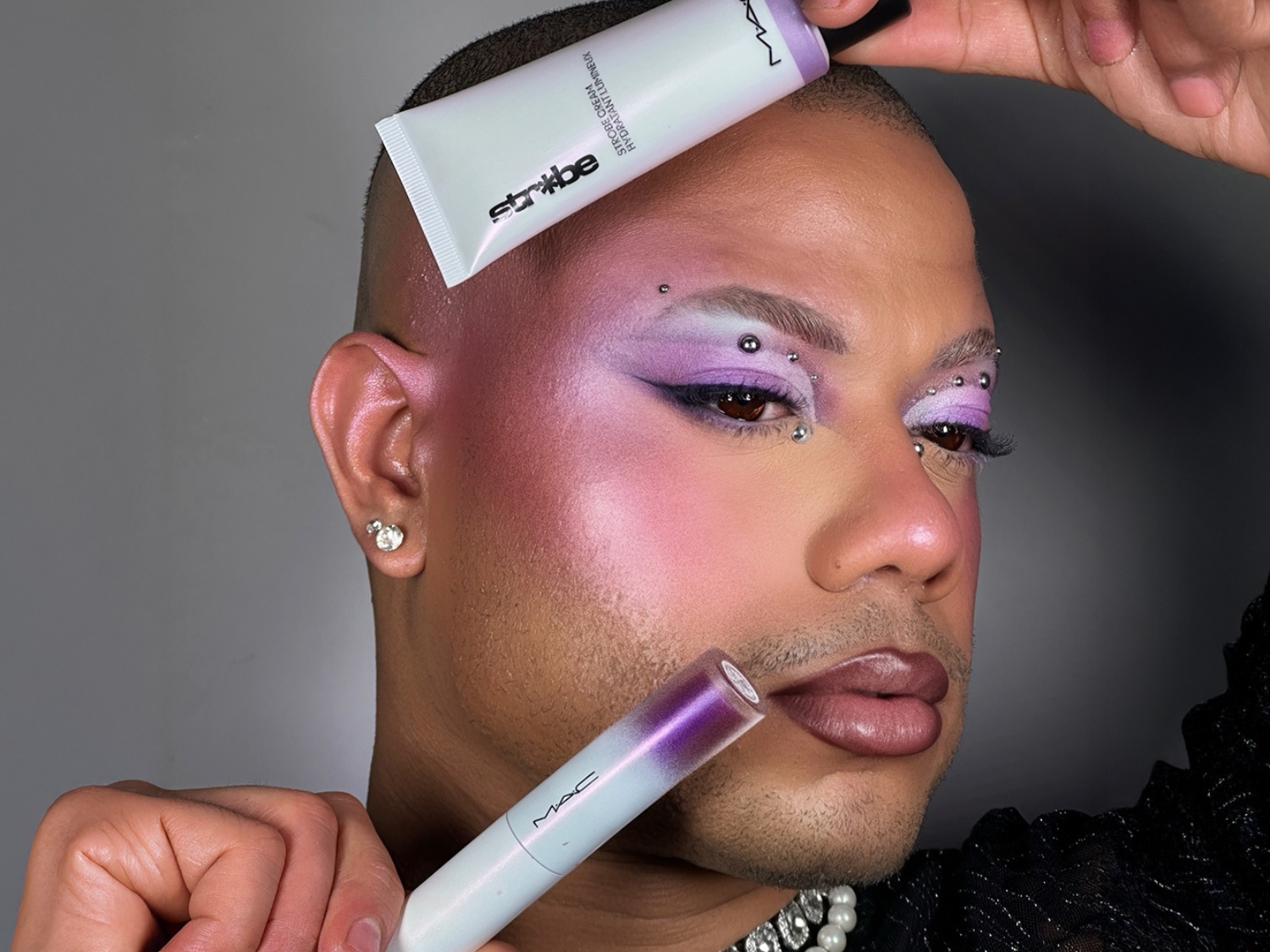 Een persoon met paarse oogschaduw en zilveren accenten, die een MAC Strobe Cream Hydratant Lumineux en een MAC lipproduct vasthoudt.