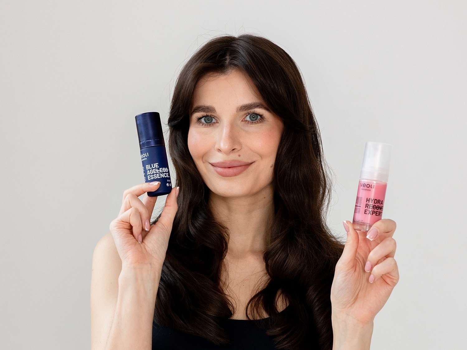 Kobieta prezentuje dwa produkty Veoli Botanica: niebieski Blue Ageless Essence i różowy Hydra Recover Expert, oba w przezroczystych butelkach z pompką.