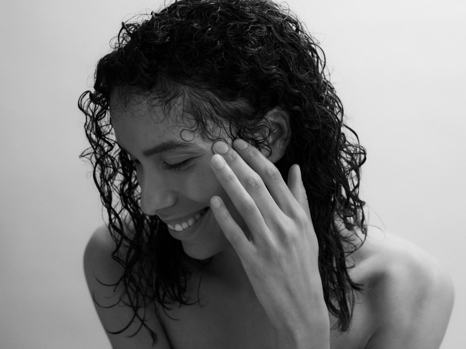 Nahaufnahme einer lächelnden Frau mit lockigem, nassem Haar, die ihr Gesicht mit der Hand berührt, in Schwarzweiß gehalten.