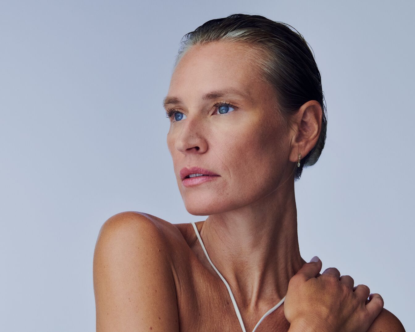 Porträt einer Frau mit hellblauen Augen, zurückgekämmtem Haar und zarten rosa Lippen, die eine Hand auf ihre Schulter legt.