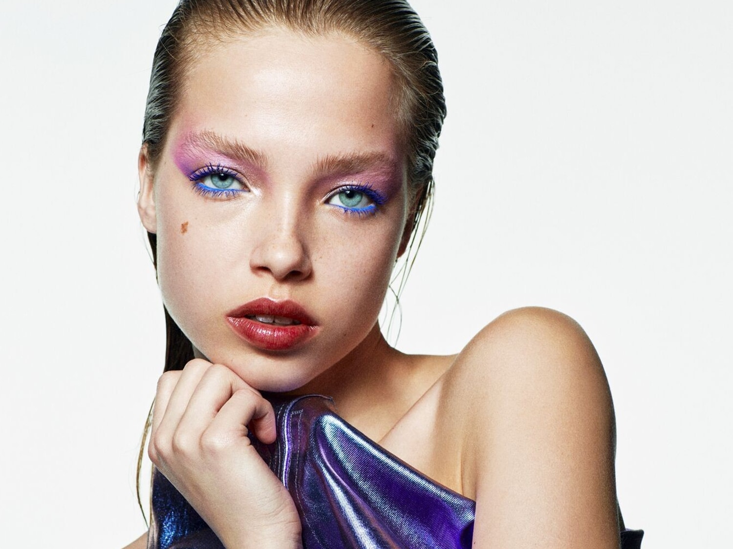 Nahaufnahme eines jungen Mädchens mit glänzendem, nass aussehendem Haar, das ein schillerndes violettes Oberteil trägt und auffälliges Make-up mit rosa Lidschatten und blauem Eyeliner.